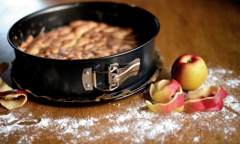 Auf dem Bild sieht man einen Kuchen in einer Kuchenspringform, vor welchem Äpfel liegen und auf der Tischplatte Mehl verstreut ist.